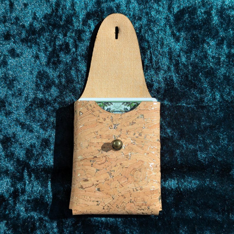 May include: A cork card holder with a light brown leather flap and a brass button closure. The interior reveals a glimpse of a patterned card. The holder is set against a dark teal background.