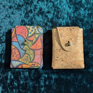 May include: Two small, rectangular wallets. One is patterned with a colourful, abstract design in shades of blue, red, yellow, and green. The other is made of light brown cork with gold flecks. Both have a flap closure with a brass-coloured button.