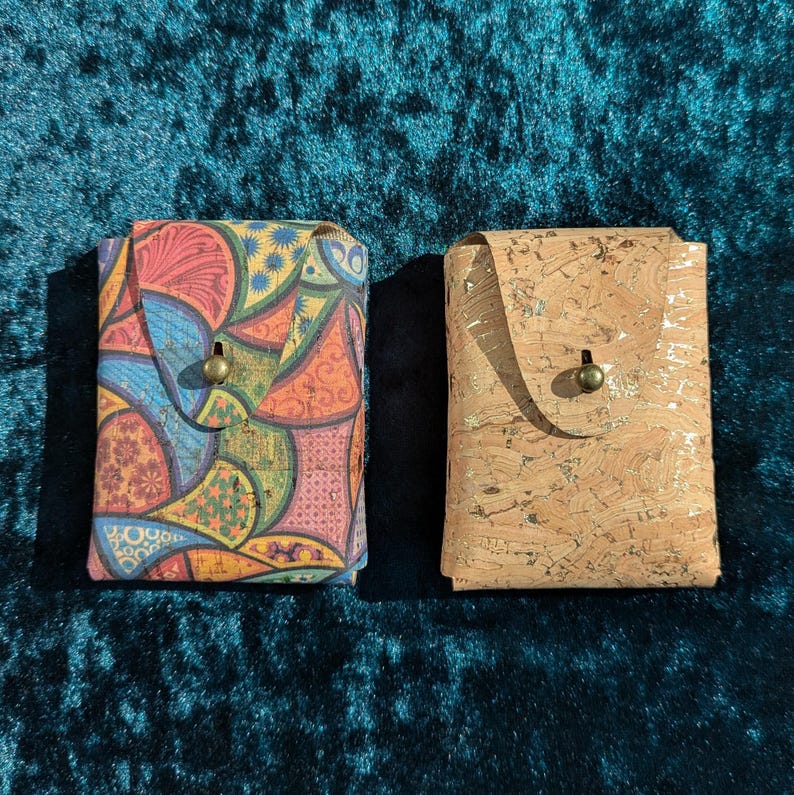 May include: Two small, rectangular wallets. One is patterned with a colourful, abstract design in shades of blue, red, yellow, and green. The other is made of light brown cork with gold flecks. Both have a flap closure with a brass-coloured button.