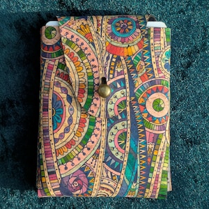 May include: A patterned cork journal in vibrant colours including orange, blue, green, and purple. The journal has a flap closure with a brass button. The cover features a detailed, symmetrical design. The journal is approximately 15 cm in height.