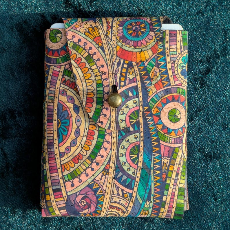 May include: A patterned cork journal in vibrant colours including orange, blue, green, and purple. The journal has a flap closure with a brass button. The cover features a detailed, symmetrical design. The journal is approximately 15 cm in height.