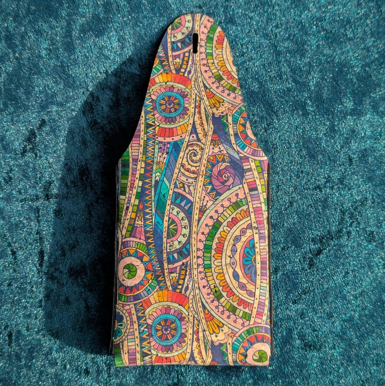 May include: A decorative beer bottle carrier with a detailed, colourful pattern. The design incorporates various shapes and colours, including blue, green, orange, pink, and yellow. It has a hole at the top for carrying.