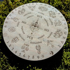 May include: A round, light-colored divination board with engraved letters, numbers, and zodiac symbols. The board features the words "yes," "no," and "unsure," along with days of the week. The center has an eye design.