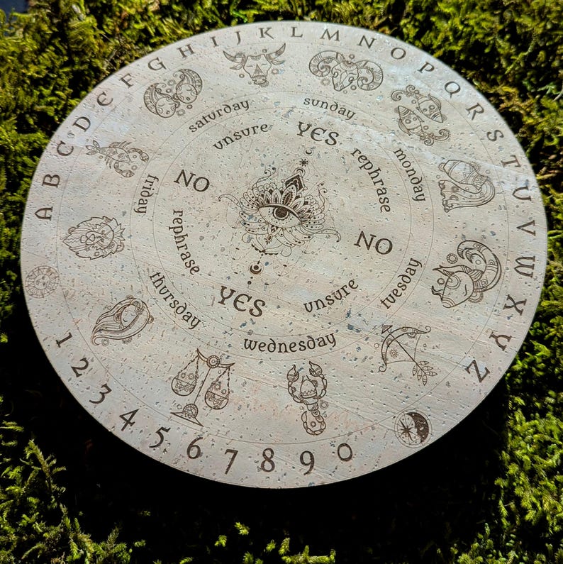 May include: A round, light-colored divination board with engraved letters, numbers, and zodiac symbols. The board features the words "yes," "no," and "unsure," along with days of the week. The center has an eye design.