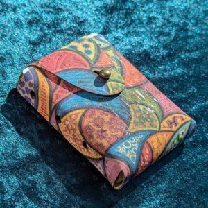May include: A colourful cork wallet with a flap closure and a brass button. The wallet features a vibrant pattern of overlapping shapes in shades of red, blue, yellow, and green. The wallet is rectangular and appears to be handmade.