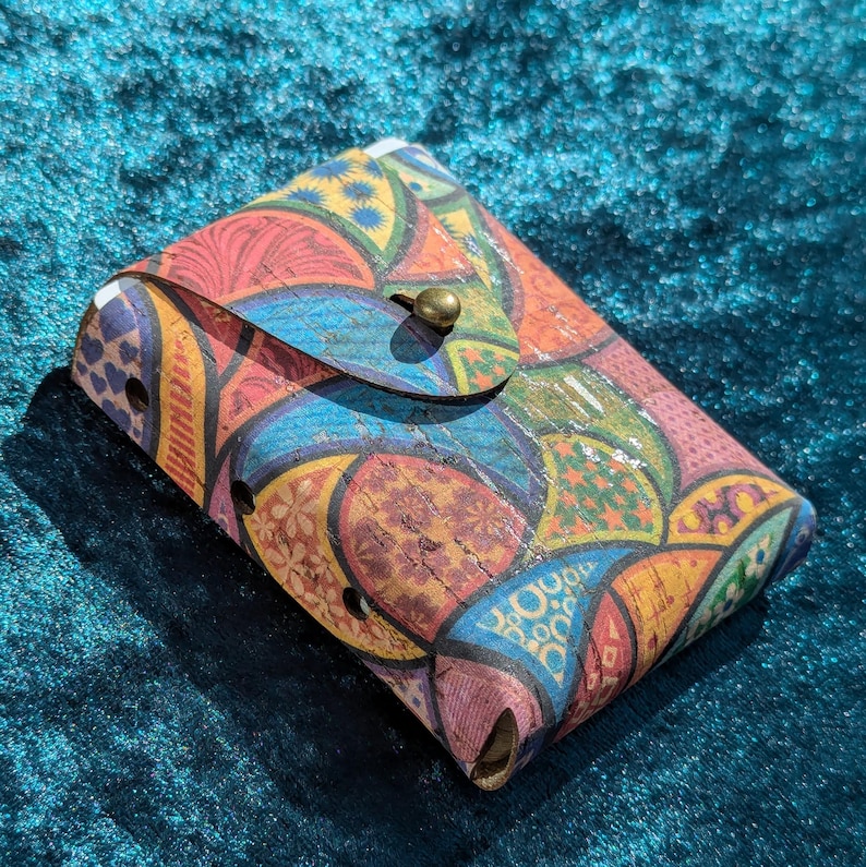 May include: A colourful cork wallet with a flap closure and a brass button. The wallet features a vibrant pattern of overlapping shapes in shades of red, blue, yellow, and green. The wallet is rectangular and appears to be handmade.