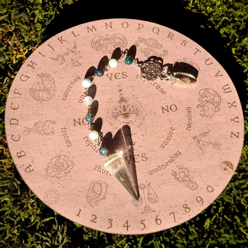 May include: A pink wooden planchette with engraved zodiac symbols, letters, numbers, and the words "yes" and "no". A pendulum with a clear crystal point and beaded chain rests on the planchette.