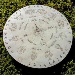 May include: A round, light-colored divination board with engraved letters, numbers, and zodiac symbols. The board features the words "yes," "no," and "rephrase," along with the days of the week. A central eye design is also present.