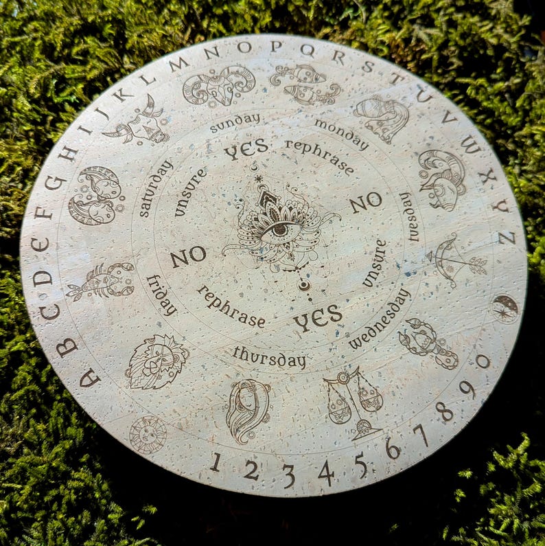 May include: A round, light-colored divination board with engraved letters, numbers, and zodiac symbols. The board features the words "yes," "no," and "rephrase," along with the days of the week. A central eye design is also present.