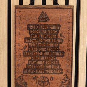 Könnte beinhalten: Ein rechteckiger Wandbehang aus braunem Holz mit einem lasergravierten Valknut-Symbol oben, flankiert von zwei Raben. Der Text lautet: PROTECT YOUR FAMILY, HONOR THE ELDERS, TEACH THE YOUNG, BE LOYAL TO YOUR FRIEND, VOICE YOUR OPINION, STAND YOUR GROUND, TAKE CHARGE WHEN OTHERS SHOW WEAKNESS, PLAY WHEN YOU CAN, WORK WHEN YOU MUST, ALWAYS LEAVE YOUR MARK. Unten sind zwei sitzende Figuren und ein Schild mit zwei Äxten abgebildet.