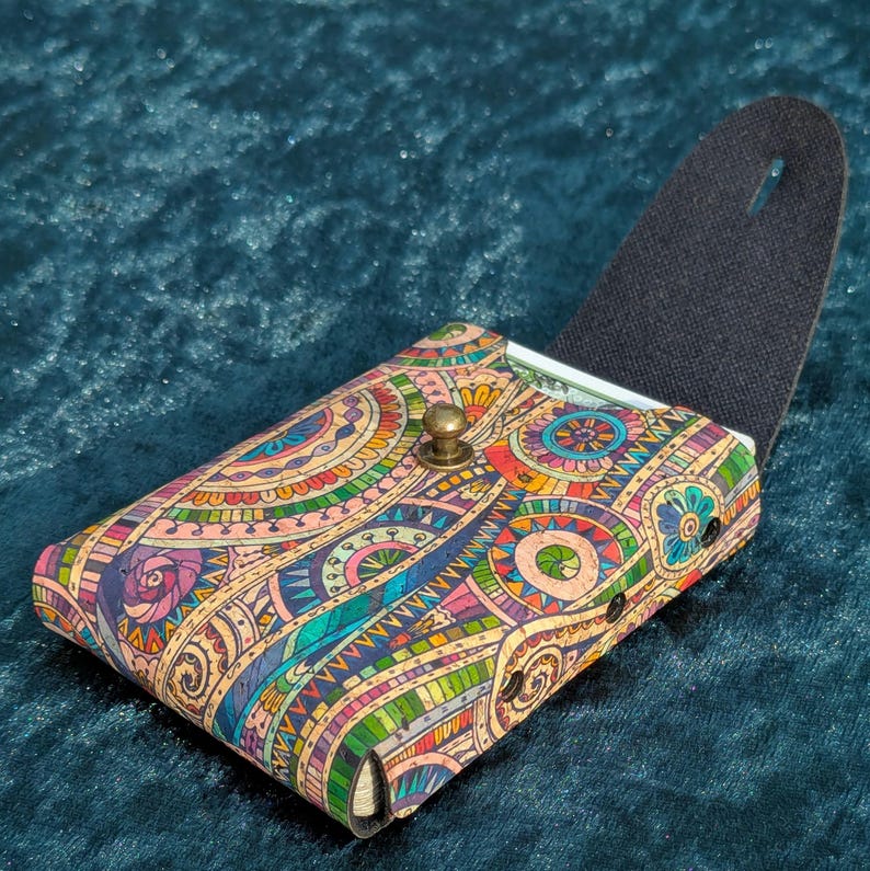 May include: A patterned wallet with an open flap, displaying a detailed design in blue, green, pink, and yellow. The wallet has a brass clasp and a black interior. The design is reminiscent of a mandala. The wallet is approximately 10 cm in length.