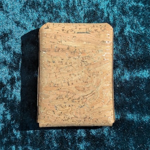 May include: A rectangular cork wallet with a natural, textured surface. The wallet is a light brown colour with darker brown edges and a subtle sheen. The background is a dark teal fabric.