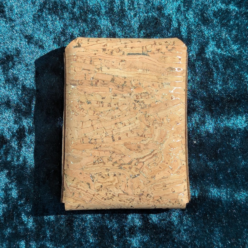 May include: A rectangular cork wallet with a natural, textured surface. The wallet is a light brown colour with darker brown edges and a subtle sheen. The background is a dark teal fabric.