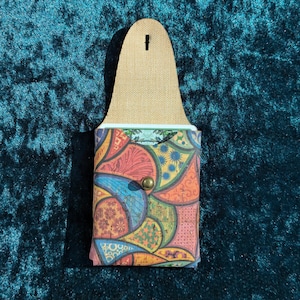May include: A colourful cork wallet with a flap and snap closure. The wallet features a vibrant patchwork design in shades of red, blue, orange, and green. The top portion is a tan leather with a hole for hanging.
