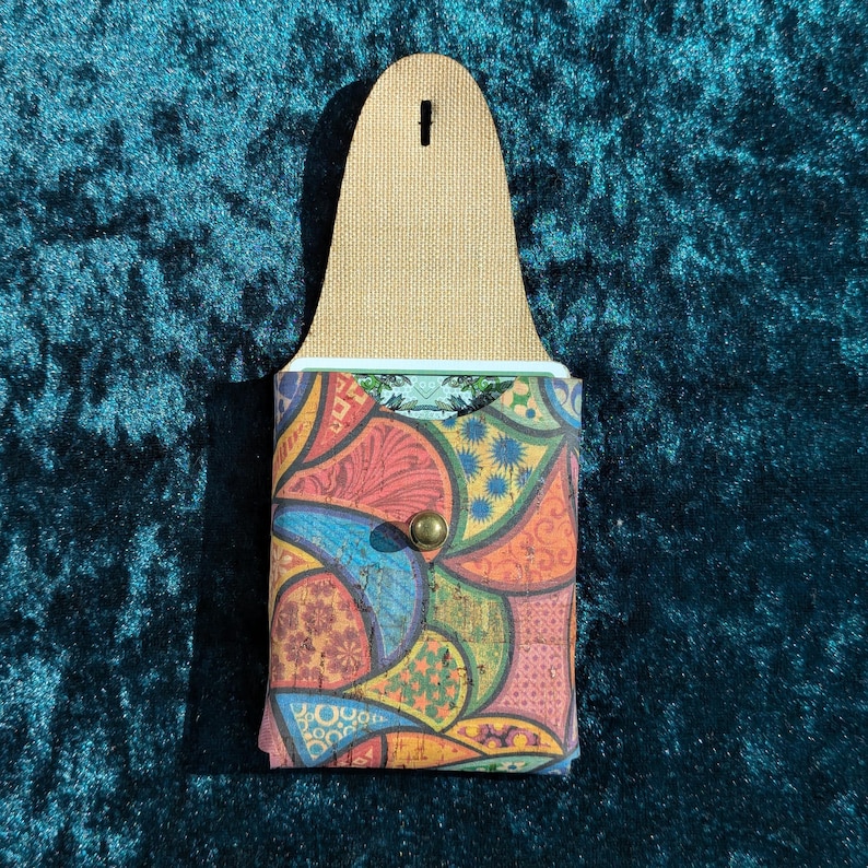 May include: A colourful cork wallet with a flap and snap closure. The wallet features a vibrant patchwork design in shades of red, blue, orange, and green. The top portion is a tan leather with a hole for hanging.