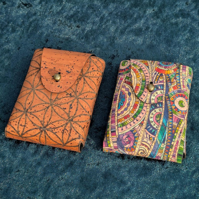 May include: Two rectangular cardholders. One is made of cork with a geometric pattern and a flap closure. The other has a colourful, intricate design with a button closure. Both are on a blue surface.