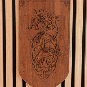Peut inclure: Une tenture murale rectangulaire en liège brun avec un bord inférieur pointu, présentant une illustration sombre gravée au laser d'un loup stylisé entrelacé de motifs de nœuds celtiques complexes. L'objet inclut un cordon noir pour suspendre.