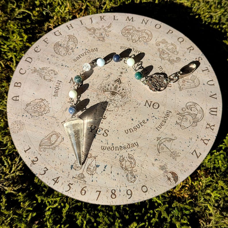 May include: A corkboard pendulum board with a crystal pendulum, a beaded chain, and a silver charm. The board features a circle of letters, numbers, and days of the week. The words "YES", "NO", and "unsure" are printed on the board.