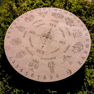 May include: Round, light-colored divination board with etched zodiac symbols, letters, and numbers. The board features the words "yes," "no," and "unsure," along with days of the week. A central eye design is surrounded by the alphabet and numbers.