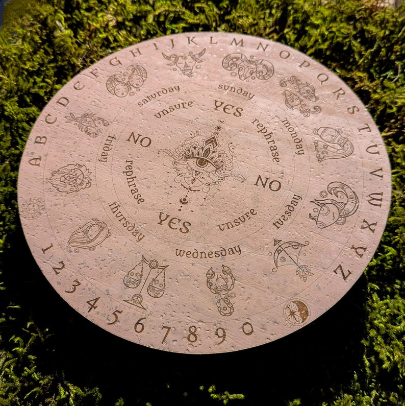 May include: Round, light-colored divination board with etched zodiac symbols, letters, and numbers. The board features the words "yes," "no," and "unsure," along with days of the week. A central eye design is surrounded by the alphabet and numbers.