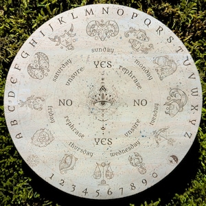 May include: A round, light-colored Ouija board with engraved letters, numbers, and zodiac symbols. The board features the words "YES," "NO," and "rephrase," along with the days of the week. The board is set against a green, mossy background.