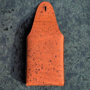 May include: An orange cork beer bottle holder with a keyhole at the top. The holder has a rectangular shape with a rounded top and is speckled with dark spots. The background is a dark blue fabric.