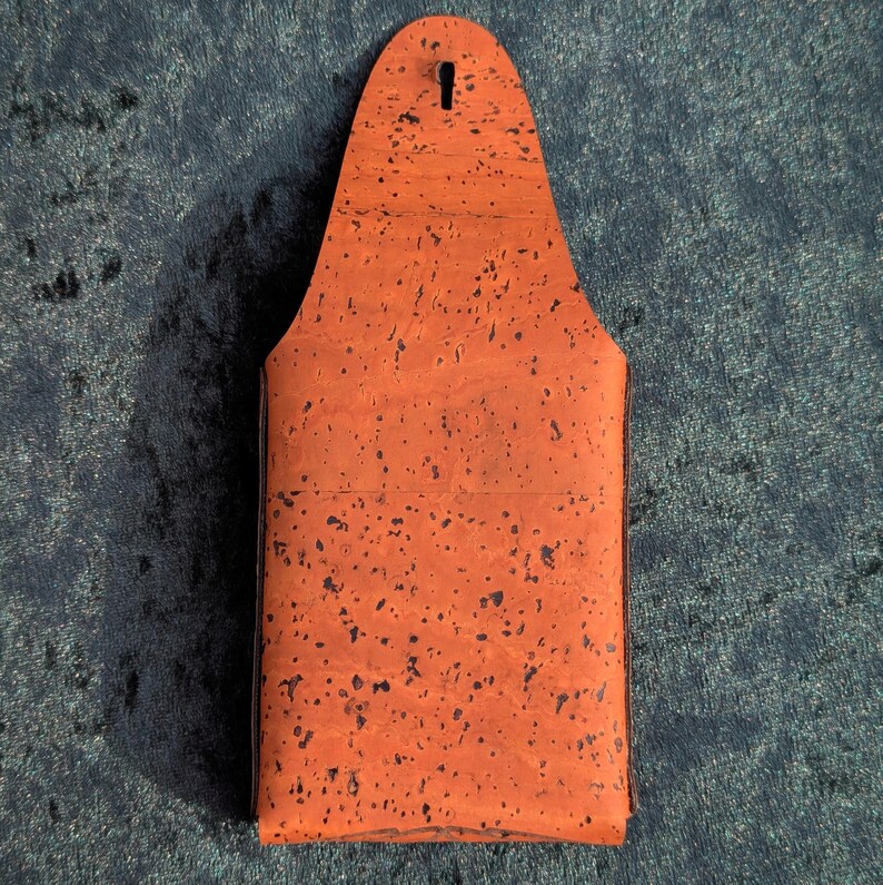May include: An orange cork beer bottle holder with a keyhole at the top. The holder has a rectangular shape with a rounded top and is speckled with dark spots. The background is a dark blue fabric.