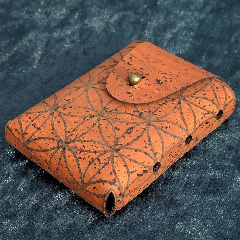 May include: An orange cork case with a geometric flower of life pattern. The case has a flap closure with a brass button and small holes on the side. The case is likely for holding small items.