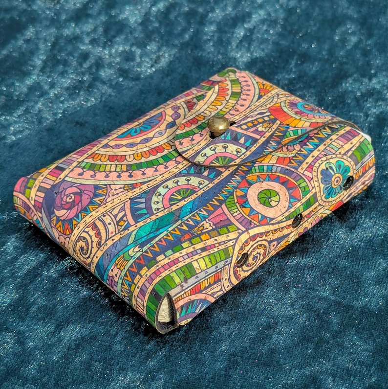 May include: A rectangular wallet with a button clasp, displaying a colourful, intricate pattern. The design features geometric shapes and floral motifs in blue, green, yellow, pink, and orange. The wallet is made from a cork-like material.