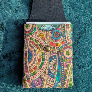 May include: A colourful cork wallet with a vibrant, patterned design featuring circles and floral elements in shades of pink, orange, blue, and green. The wallet has a black strap and a brass button closure. The wallet is designed to hold cards.