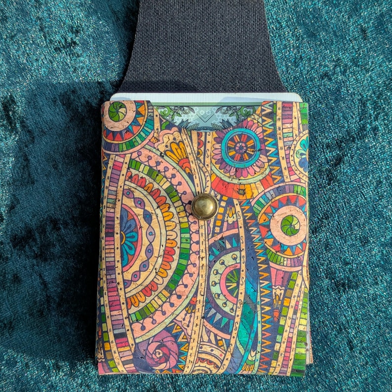 May include: A colourful cork wallet with a vibrant, patterned design featuring circles and floral elements in shades of pink, orange, blue, and green. The wallet has a black strap and a brass button closure. The wallet is designed to hold cards.