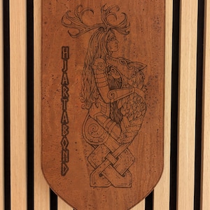 Könnte beinhalten: Ein rechteckiger Wandbehang aus Holz mit spitz zulaufender Unterkante, der eine lasergravierte Illustration einer Figur mit Geweih zeigt, die einen Wolf umarmt. Der Text "HJARTABOND" ist vertikal auf der linken Seite eingraviert.