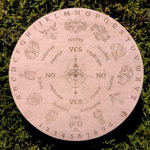 May include: A round, light pink divination board with engraved zodiac symbols, letters, numbers, and the words "yes," "no," and "rephrase." The board features a central eye design and is surrounded by the alphabet and numbers.