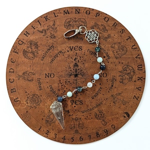 Pode incluir: Um pêndulo com uma ponta de cristal transparente facetada presa a uma corrente prateada com contas alternadas em azul escuro, azul claro e transparentes, finalizado com um fecho lagosta de metal e um pingente de flor da vida.