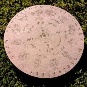 May include: A round, light-colored wooden Ouija board with engraved letters, numbers, and zodiac symbols. The board features the words "yes," "no," and "rephrase," along with the days of the week. A central eye design is also present.