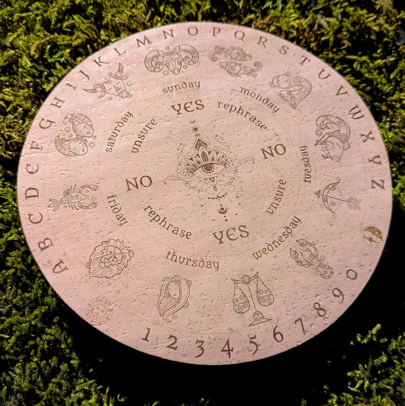 May include: A round, light-colored wooden Ouija board with engraved letters, numbers, and zodiac symbols. The board features the words "yes," "no," and "rephrase," along with the days of the week. A central eye design is also present.
