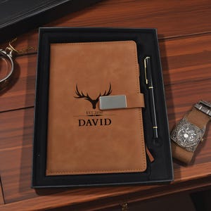 May include: A brown leather journal with a pen, presented in a black box. The journal features a deer antler logo, the text "EST. 2030", and the name "DAVID". A watch is visible on the right.