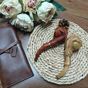 May include: Two carved wooden tobacco pipes with curved stems sit on a woven mat. One pipe is dark brown with a carved design, the other is light brown with a bamboo-like stem. A brown leather case is on the left.