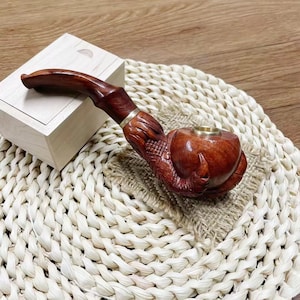 May include: A wooden smoking pipe with a detailed carving of a claw. The pipe is brown and has a brass band around the stem. The pipe is sitting on a woven mat.
