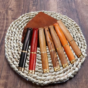 Puede incluir: Siete bolígrafos de madera con detalles dorados, dispuestos en círculo sobre una estera de paja tejida. Los bolígrafos son de varios tonos de marrón, rojo y negro.