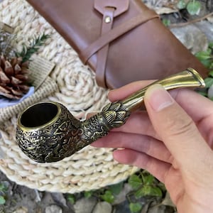 May include: A gold-colored metal pipe with intricate floral designs. The pipe has a round bowl and a long, curved stem.