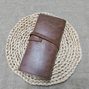 May include: A brown leather wallet with a strap closure. The wallet is rectangular and has a textured surface. The wallet is placed on a woven, circular placemat.