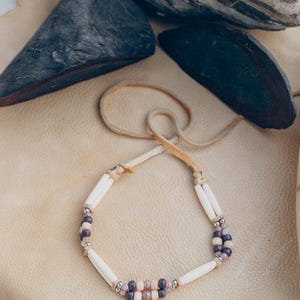 Può includere: Una collana in cordoncino di cuoio bianco con perline di osso bianco e perline di vetro viola e rosa.