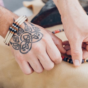 May include: A person's hand with a Celtic knot tattoo is wearing a layered bone bracelet and holding a multi-coloured beaded necklace.