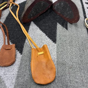 May include: Several small leather pouches in shades of brown and yellow, each with a long strap. The pouches are on a grey and black patterned textile. Also visible are two dark, rounded stones and a woven ring.