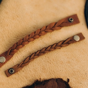 May include: Two brown leather braided bracelets with silver metal clasps. The bracelets are lying on a light brown surface.
