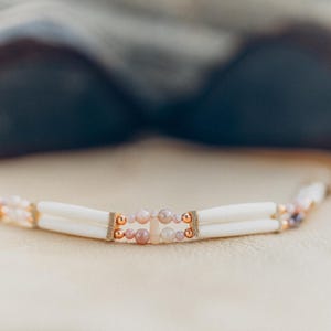 Può includere: Una collana di perline bianche con accenti rosa e dorati. La collana è realizzata con perline di osso e presenta un gruppo centrale di pietre rosa.