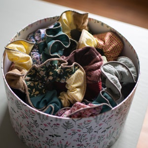 Peut inclure: Une boîte blanche à motif floral remplie de chouchous en tissu colorés. Les chouchous sont faits de différents tissus, dont du plaid, du floral et des couleurs unies.