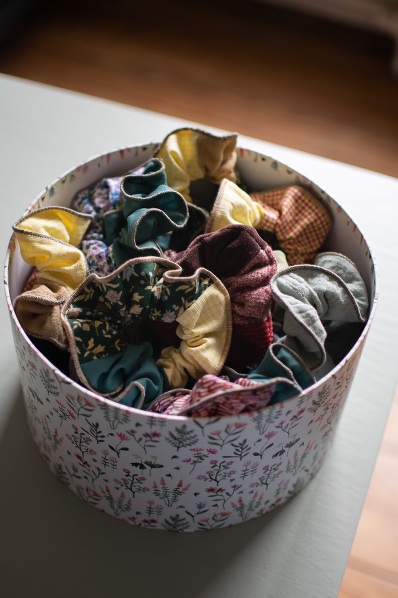 May include: A white box with a floral pattern filled with colorful fabric scrunchies. The scrunchies are made of different fabrics, including plaid, floral, and solid colors.