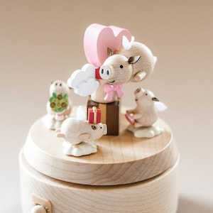 May include: A wooden music box with a pink heart and four pig figurines. The pig figurines are painted white and have wings. The music box is made of light brown wood and has a small knob on the side. The pig figurines are arranged around the heart, and the music box is on a light brown surface.